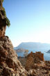 Kalymnos - The remaining structures of the Kastri under the towering cliffs which rise protectively behind; Tendelos Island in the background